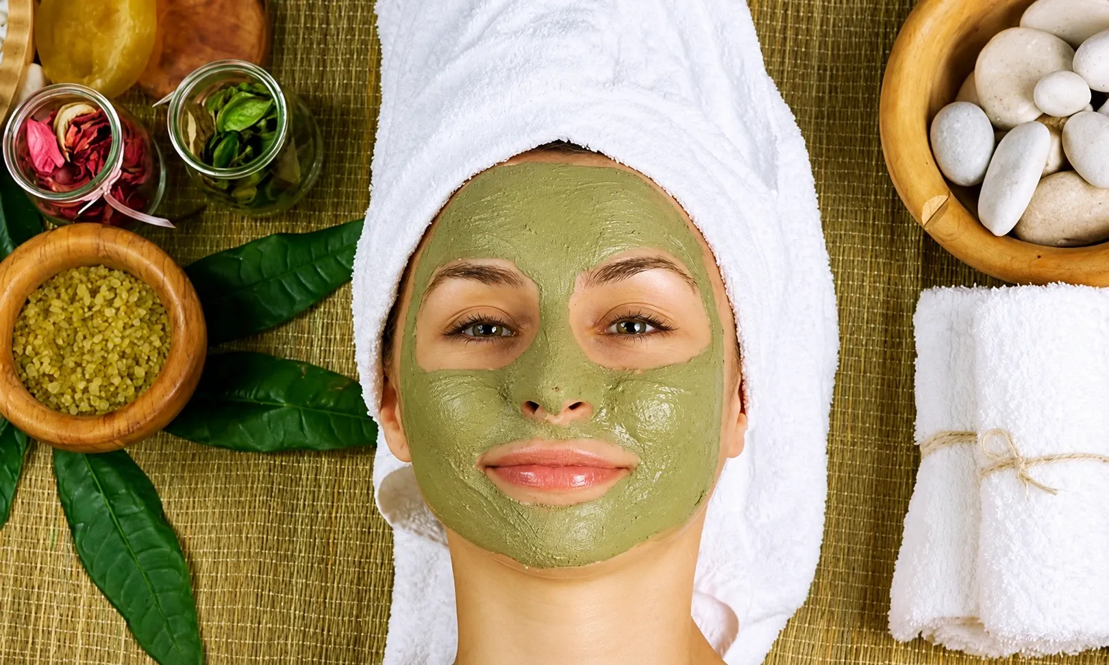 guest wearing a green herbal face mask, with bowls of herbs and petals around her